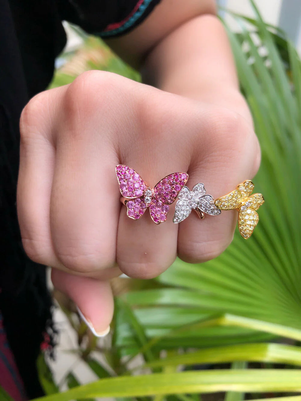Three butterfly-shaped rings in pink, silver, and gold on a finger with a blurred green leafy background.