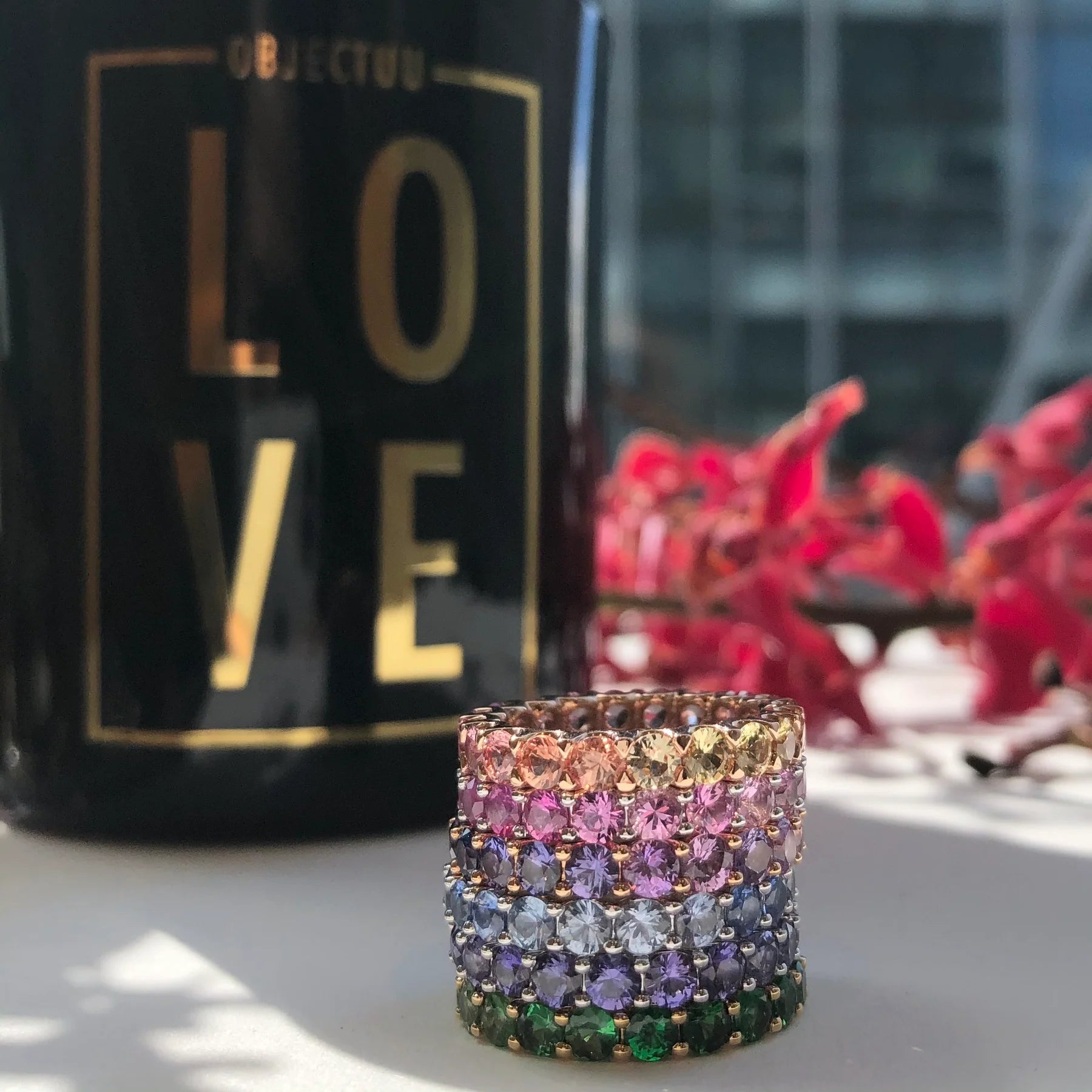Multicolored beaded ring on a reflective surface with a blurred 'LOVE' bottle in the background.