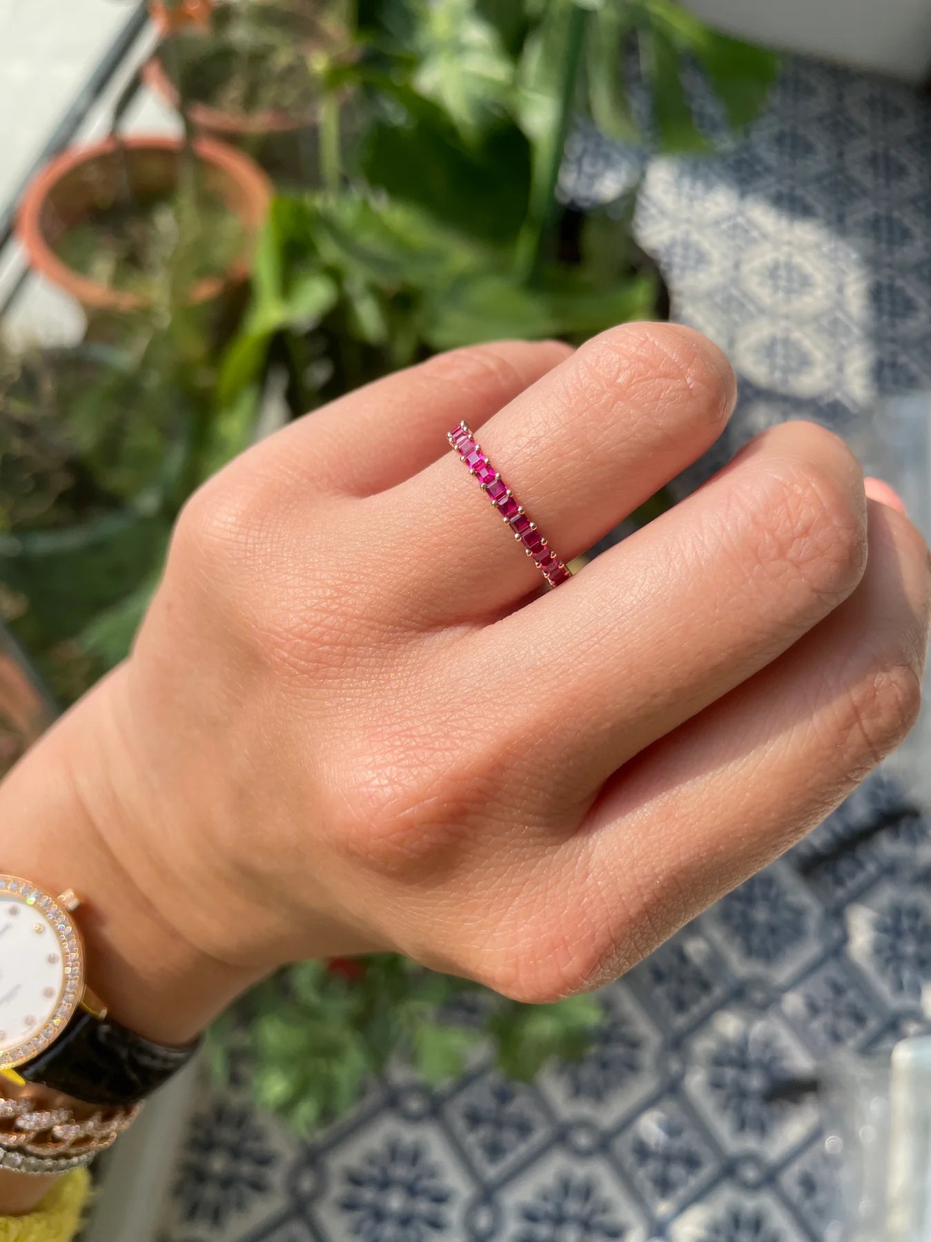 Hand wearing a pink ring with a blurred background of plants and a patterned surface.