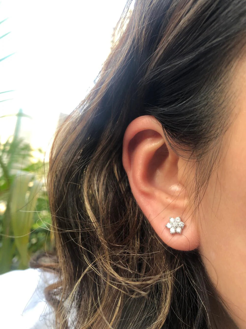 Close-up of an ear wearing a diamond flower earring with blurred greenery in the background