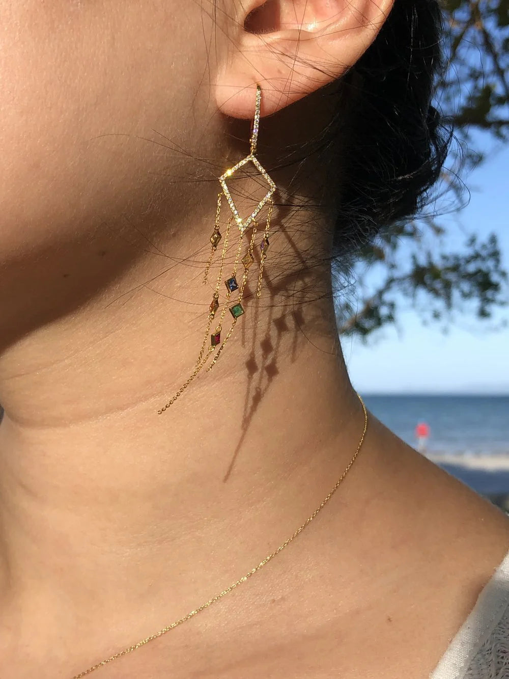 Close-up of a person wearing gold earrings with gemstones against a blurred outdoor background.